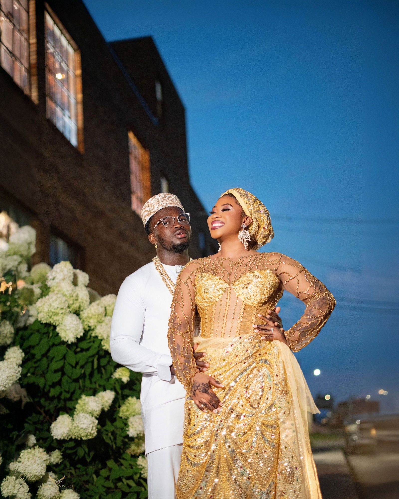 Couple in gold traditional attire at dusk, beside white hydrangeas