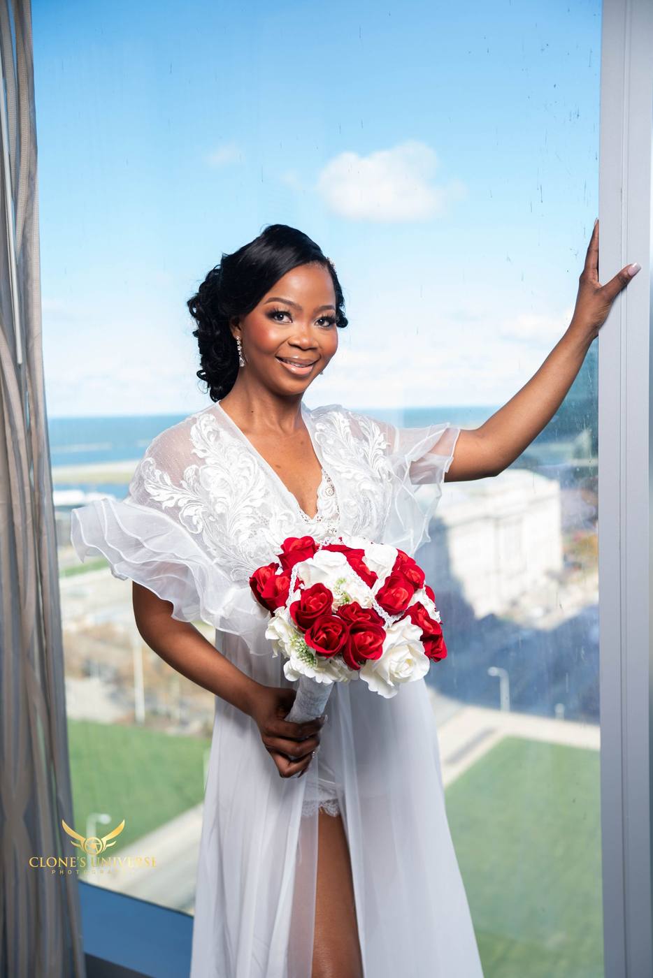 Bride in a lace robe holding red and white roses at a high-rise window