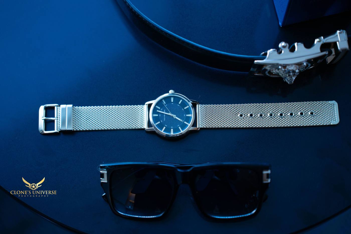 Groom’s watch, sunglasses and belt laid out on dark blue