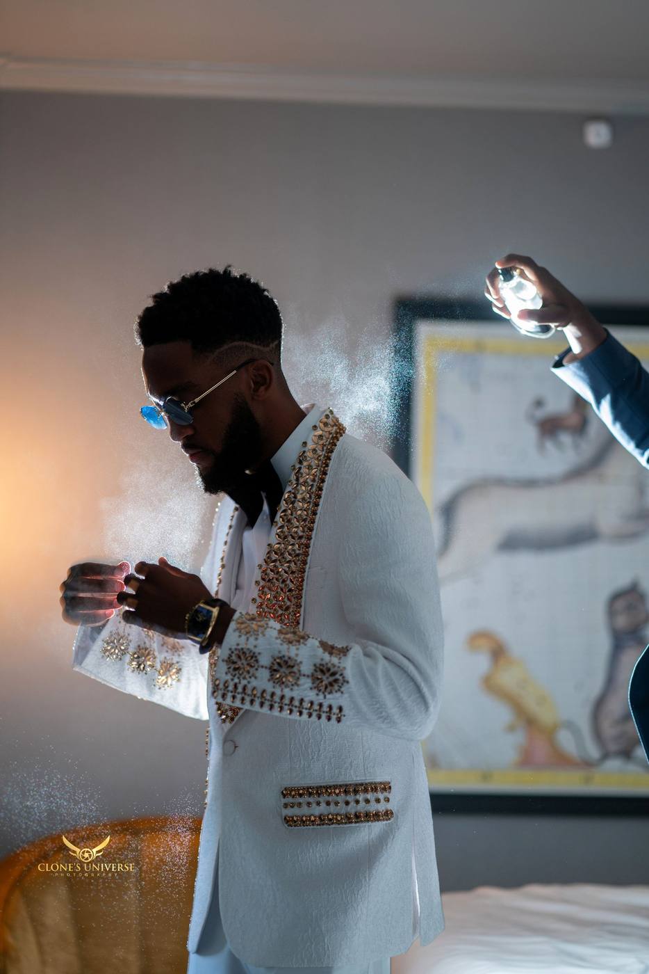 Groom in a white jacket with gold beading being spritzed with cologne